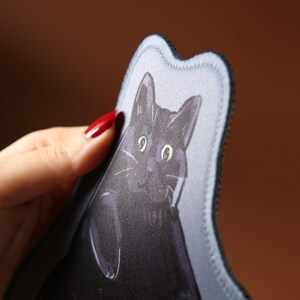 May include: A fabric item featuring a black cat illustration. The cat has large eyes and a detailed face. The item has a wavy edge and a black border. The background is blurred, with a brown and green hue.