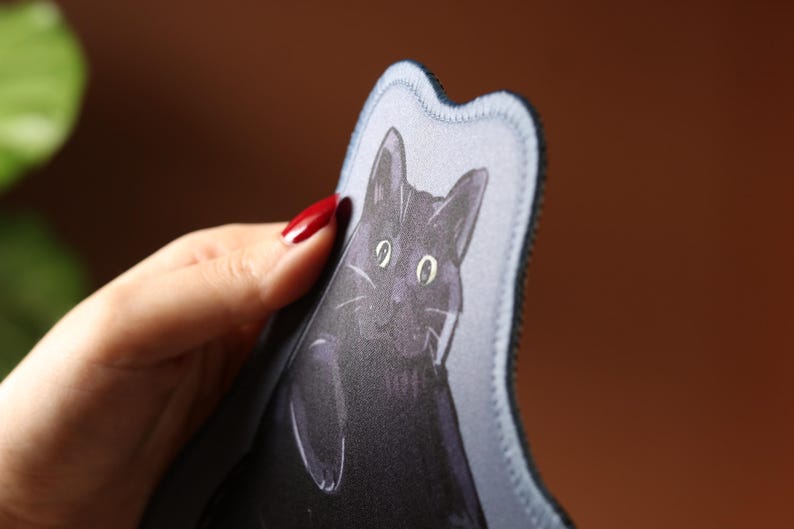 May include: A fabric item featuring a black cat illustration. The cat has large eyes and a detailed face. The item has a wavy edge and a black border. The background is blurred, with a brown and green hue.