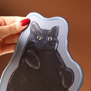 May include: A black cat illustration on a light blue, irregularly shaped patch. The cat has wide eyes and a relaxed pose. The patch has a stitched border and is held up by a hand with red nail polish.