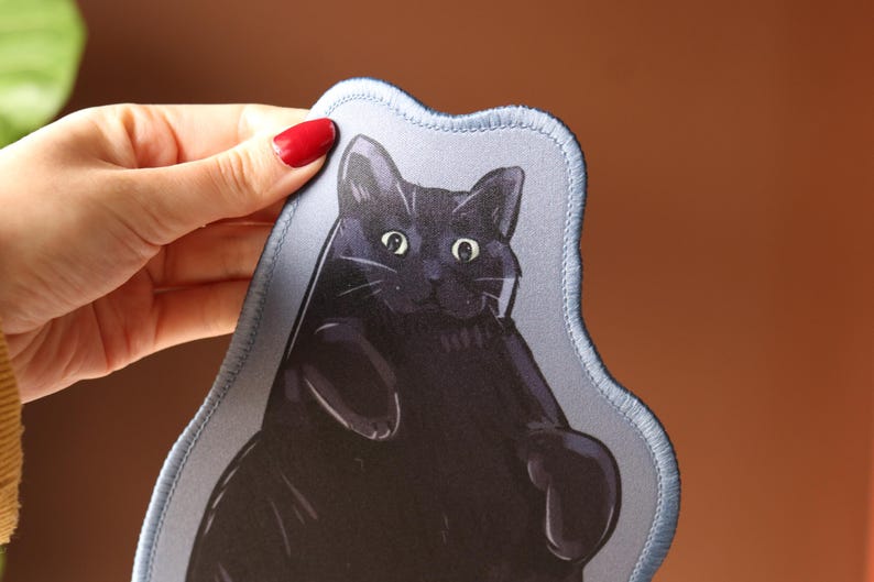May include: A black cat illustration on a light blue, irregularly shaped patch. The cat has wide eyes and a relaxed pose. The patch has a stitched border and is held up by a hand with red nail polish.