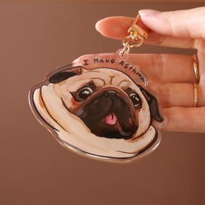 May include: A clear acrylic pug dog keychain with a gold-coloured clasp. The pug has a tan and black face with a pink tongue. The keychain has the text "I Have Asthma" on a banner above the dog's head.