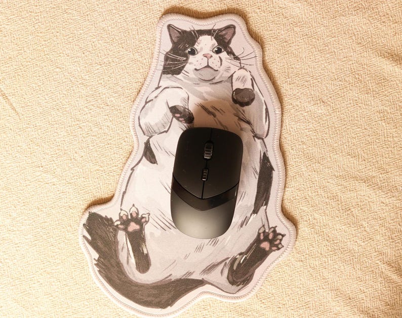May include: A white and black cat mousepad with a cartoon cat lying on its back. A black computer mouse is on top of the cat.