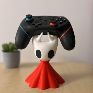 May include: A black games controller sits on a white and red decorative stand. The controller has blue and orange accents. The stand features a white head with black eyes and a red cape, resembling a video game character.