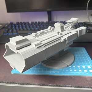 Puede incluir: Un modelo de nave espacial impreso en 3D, pintado en color gris claro. La nave espacial tiene un cuerpo largo y estrecho con una nariz puntiaguda y un ala grande y plana a cada lado. El modelo est&aacute; sentado en una estera de corte azul y blanca.