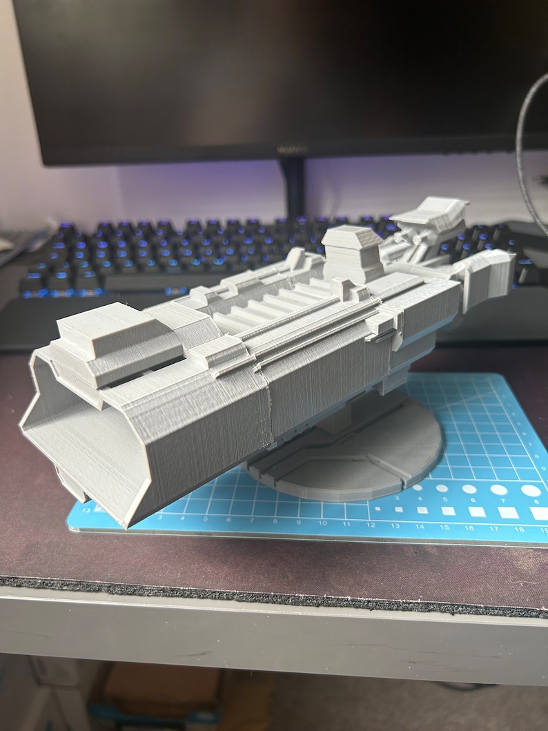 Puede incluir: Un modelo de nave espacial impreso en 3D, pintado en color gris claro. La nave espacial tiene un cuerpo largo y estrecho con una nariz puntiaguda y un ala grande y plana a cada lado. El modelo est&aacute; sentado en una estera de corte azul y blanca.
