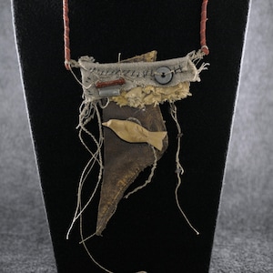 May include: A mixed-media necklace featuring a brown leather base with a beige fabric top layer, a small wooden fish cutout, a dark button, and a red cylindrical bead. The pendant hangs from a thin, reddish-brown cord with visible frayed threads.