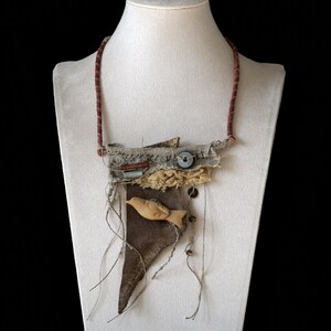 May include: A mixed-media necklace featuring a dark brown leather triangular base, a carved light-colored wooden bird, frayed beige fabric layers, a dark button, and visible stitching. The necklace hangs from a thin, textured reddish-brown cord.