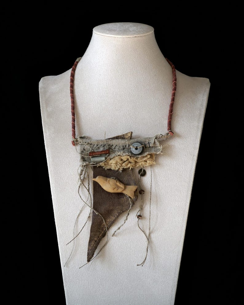 May include: A mixed-media necklace featuring a dark brown leather triangular base, a carved light-colored wooden bird, frayed beige fabric layers, a dark button, and visible stitching. The necklace hangs from a thin, textured reddish-brown cord.