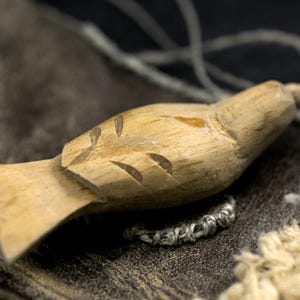 May include: A light brown, hand-carved wooden bird figurine featuring simple incised lines to represent wings and feathers. The small, smooth wooden sculpture has a pointed beak and a flat, fan-shaped tail, showcasing natural wood grain textures.