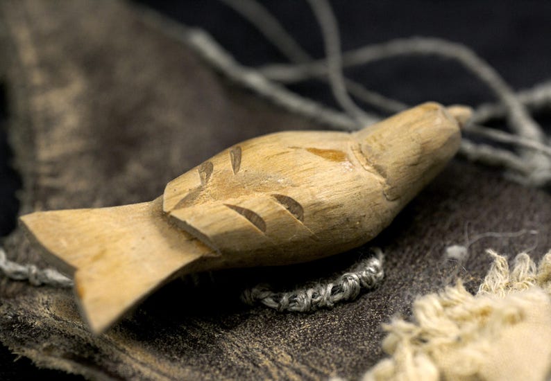 May include: A light brown, hand-carved wooden bird figurine featuring simple incised lines to represent wings and feathers. The small, smooth wooden sculpture has a pointed beak and a flat, fan-shaped tail, showcasing natural wood grain textures.