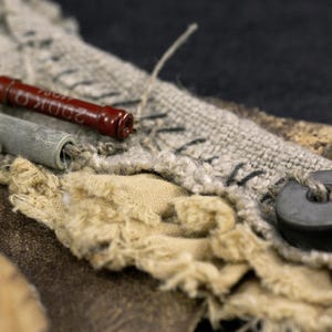 May include: A textured composition featuring a dark red cylindrical electronic resistor with 200K ohm text, a grey cylindrical component, a black circular bead, and layered beige and grey fabric scraps with visible black stitching and frayed edges.
