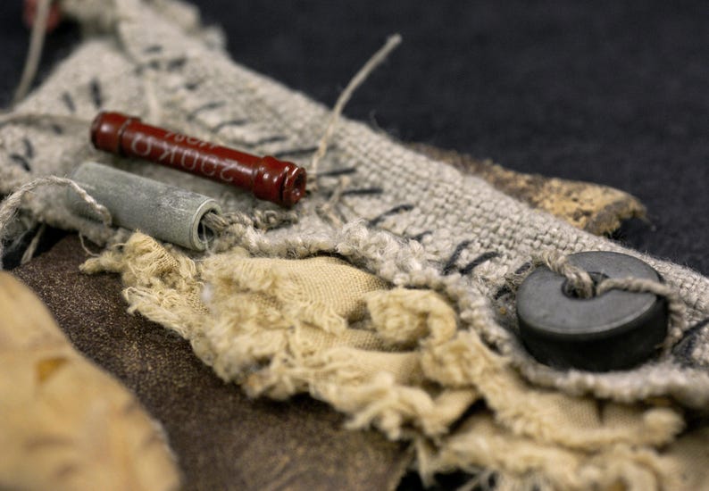 May include: A textured composition featuring a dark red cylindrical electronic resistor with 200K ohm text, a grey cylindrical component, a black circular bead, and layered beige and grey fabric scraps with visible black stitching and frayed edges.