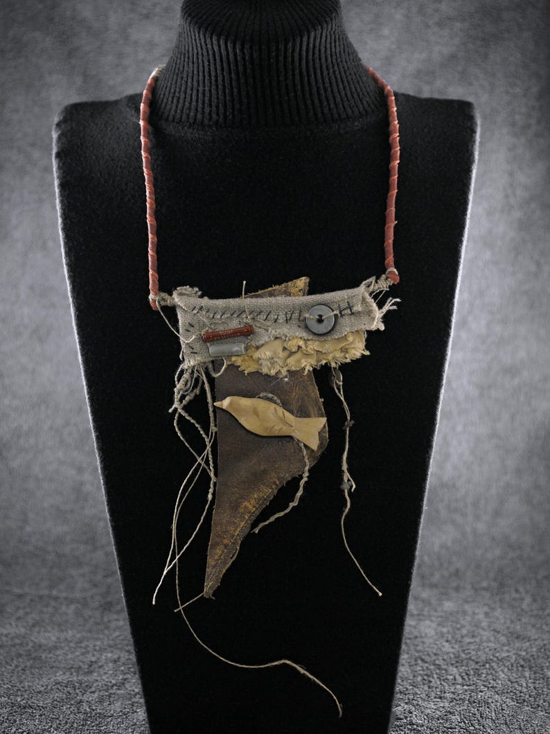 May include: A mixed-media necklace featuring a brown leather base with a beige fabric top layer, a small wooden fish cutout, a dark button, and a red cylindrical bead. The pendant hangs from a thin, reddish-brown cord with visible frayed threads.