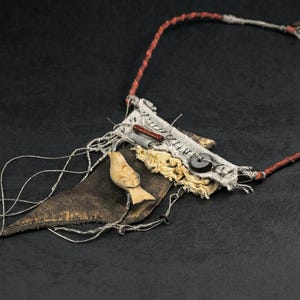 May include: A necklace featuring a dark brown leather triangle, a light-colored carved wooden bird, and a rectangular fabric patch with visible stitching. The piece includes a black button, a small red cylinder, and a thin red and grey cord.