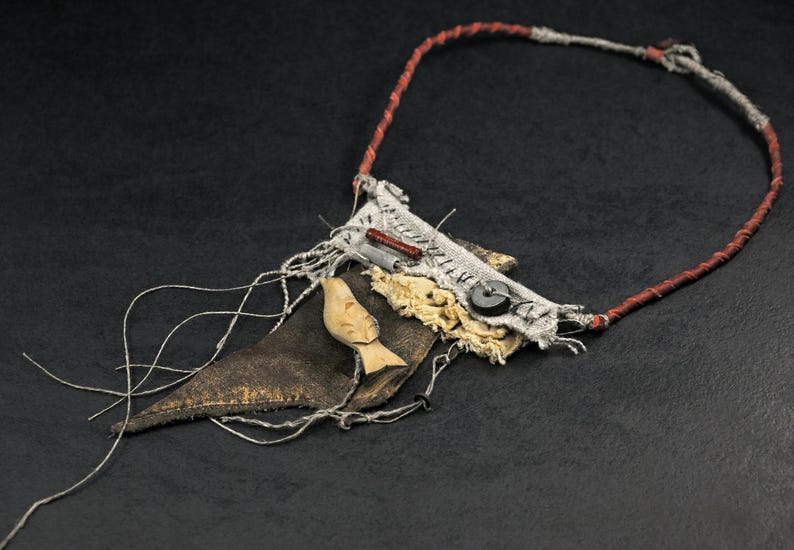 May include: A necklace featuring a dark brown leather triangle, a light-colored carved wooden bird, and a rectangular fabric patch with visible stitching. The piece includes a black button, a small red cylinder, and a thin red and grey cord.