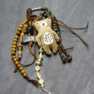 May include: A brown and white teddy bear charm with a beaded necklace, a leather cord, and a silver clip. The charm is attached to a brown and white rope.