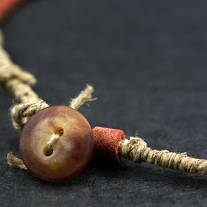 May include: A necklace featuring a brown two-hole button closure, a small cylindrical red bead, and a tan twine cord. The cord is wrapped with thin fibers and knotted to secure the button, creating a functional fastener for the jewelry piece.