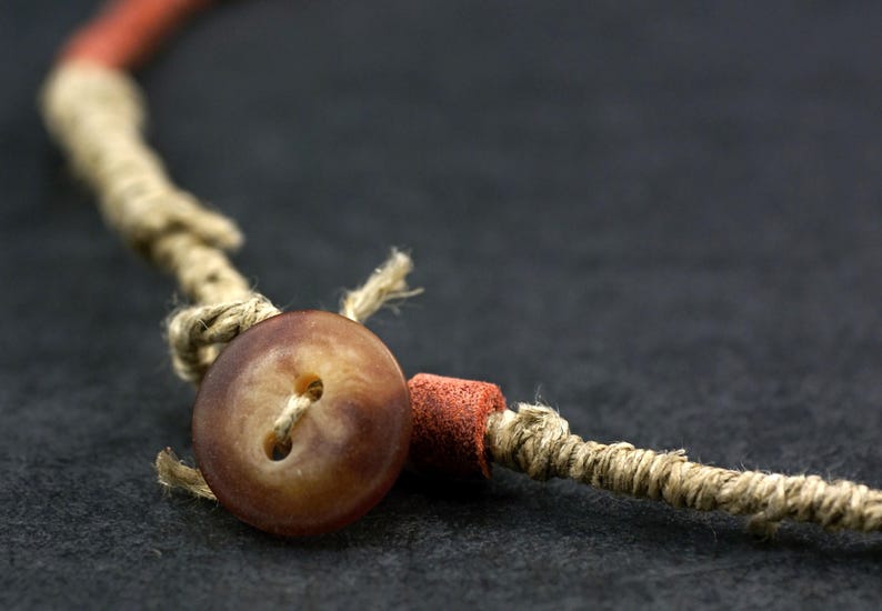 May include: A necklace featuring a brown two-hole button closure, a small cylindrical red bead, and a tan twine cord. The cord is wrapped with thin fibers and knotted to secure the button, creating a functional fastener for the jewelry piece.