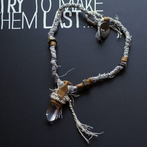May include: A gray and white fabric necklace with brown leather accents and a clear crystal pendant. The necklace has a heart shape.