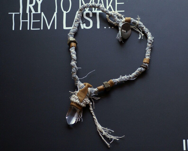 May include: A gray and white fabric necklace with brown leather accents and a clear crystal pendant. The necklace has a heart shape.