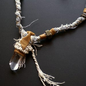 May include: A crystal pendant necklace with a clear quartz crystal, wrapped in brown leather and white fabric, and a silver metal accent. The necklace is made with a gray and white braided cord.