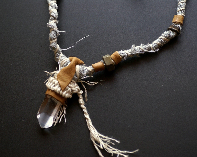 May include: A crystal pendant necklace with a clear quartz crystal, wrapped in brown leather and white fabric, and a silver metal accent. The necklace is made with a gray and white braided cord.