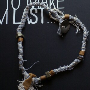 May include: A gray and white fabric rope necklace with metal accents and a button. The rope is tied in a heart shape.