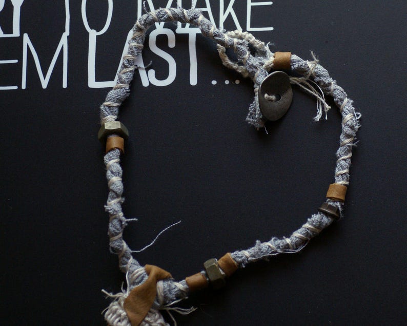 May include: A gray and white fabric rope necklace with metal accents and a button. The rope is tied in a heart shape.