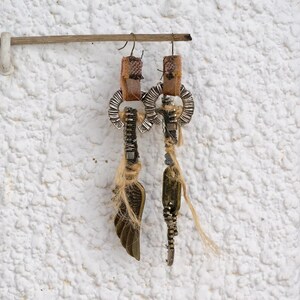 May include: A pair of handmade earrings with a rustic, bohemian style. Each earring features a silver-toned metal charm with a wing design, a brown leather tassel, and a silver-toned metal hoop. The earrings are hanging from a wooden dowel.