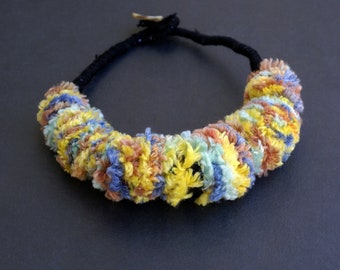 May include: A colorful necklace made of yarn with a black cord. The yarn is in shades of blue, yellow, and pink.