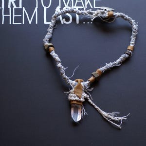 May include: A gray and brown fabric necklace with a crystal pendant. The necklace has a tassel and is tied with a knot.