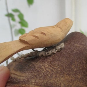May include: A light brown wooden bird carving featuring simple, hand-carved wing details and a flat tail. The bird is attached to a dark brown, textured, rounded object with a piece of light tan woven twine visible underneath the wooden figure.