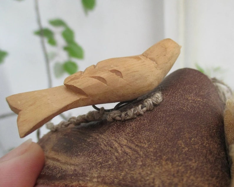 May include: A light brown wooden bird carving featuring simple, hand-carved wing details and a flat tail. The bird is attached to a dark brown, textured, rounded object with a piece of light tan woven twine visible underneath the wooden figure.