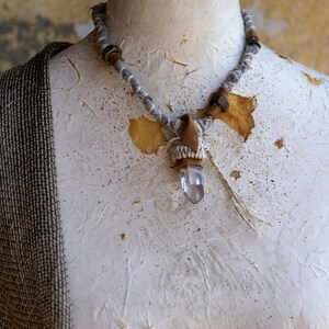 May include: A white crystal pendant necklace with a brown and white fabric wrap and brown beads. The necklace is on a white mannequin.