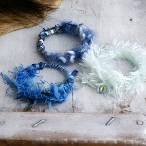 Könnte beinhalten: Drei handgefertigte Armbänder aus blauem und weißem Garn. Die Armbänder liegen auf einer weißen Holzoberfläche.