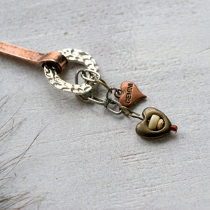 Puede incluir: Un collar con un dije de cobre y plata, con un dije en forma de corazón que dice "Géminis" y un dije redondo con una superficie texturizada.