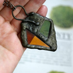May include: A handmade pendant necklace with a geometric design. The pendant features a dark metal frame with three sections of colored glass: green, orange, and amber. The necklace has a chain and a textured, organic appearance.