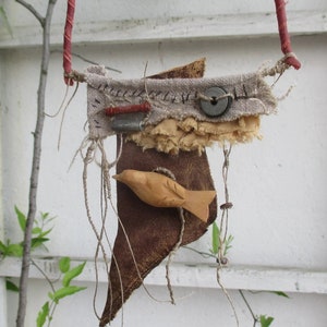 May include: A mixed media wall hanging featuring a carved wooden bird attached to a dark brown leather piece. Above it, a beige fabric strip is adorned with a grey button, a small red cylinder, a metallic tube, and black hand-stitched details.