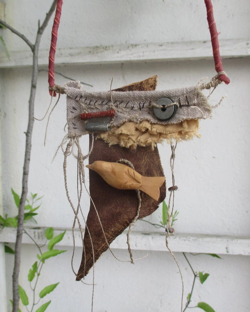 May include: A mixed media wall hanging featuring a carved wooden bird attached to a dark brown leather piece. Above it, a beige fabric strip is adorned with a grey button, a small red cylinder, a metallic tube, and black hand-stitched details.