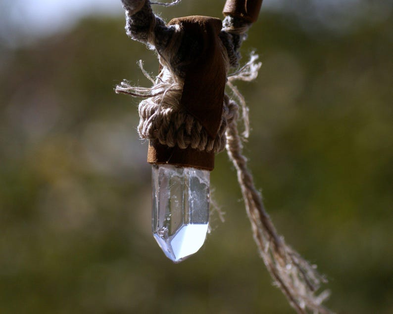 May include: A clear quartz crystal pendant hanging from a brown leather cord with a knotted rope detail.
