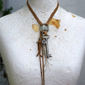 Puede incluir: Un collar de cuero marrón con un dije de llave de plata y un dije de plata con diamantes de imitación. El collar está colgado de un maniquí blanco.