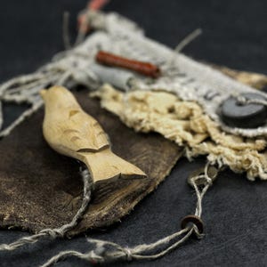 May include: A carved light-colored wooden bird figurine resting on a dark brown piece of leather. The composition includes frayed off-white fabric, a small dark cylindrical bead, a flat black circular button, and thin light-colored twine.