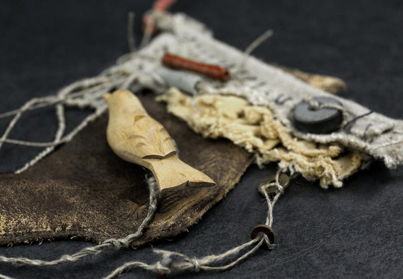 May include: A carved light-colored wooden bird figurine resting on a dark brown piece of leather. The composition includes frayed off-white fabric, a small dark cylindrical bead, a flat black circular button, and thin light-colored twine.