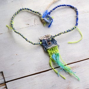 Puede incluir: Un collar de hilo azul, verde y blanco con un dije de bolsillo de mezclilla. El collar está hecho de varios hilos de hilo y tiene un borde deshilachado.