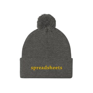 Peut inclure: Un bonnet en tricot gris avec un pompon moelleux sur le dessus. Le bonnet a le mot "spreadsheets" brodé sur le devant en jaune.