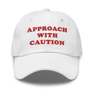 Puede incluir: Gorra de béisbol blanca con la frase bordada en rojo "APPROACH WITH CAUTION". La gorra tiene una visera curva y un botón en la parte superior. Un accesorio casual.
