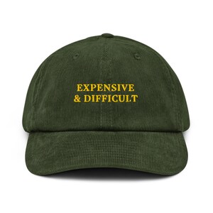 Peut inclure: Une casquette de baseball en velours côtelé vert foncé avec le texte "EXPENSIVE & DIFFICULT" brodé en jaune.