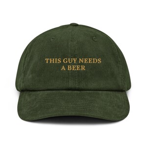 May include: A green corduroy baseball cap with the text "This Guy Needs A Beer" embroidered in gold.
