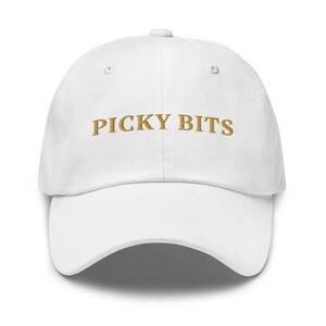 Pode incluir: Boné de beisebol branco com aba curva. O texto "PICKY BITS" é bordado em dourado na frente. O boné apresenta um design clássico de seis painéis, adequado para várias roupas casuais.
