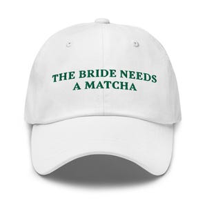 May include: White baseball cap with the text "THE BRIDE NEEDS A MATCHA" in green lettering. The cap has a curved brim and a button on top. A casual accessory for a bride.